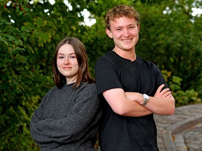 Zwei junge Menschen stehen vor einem natürlichen Hintergrund aus Blättern und schauen in die Kamera.